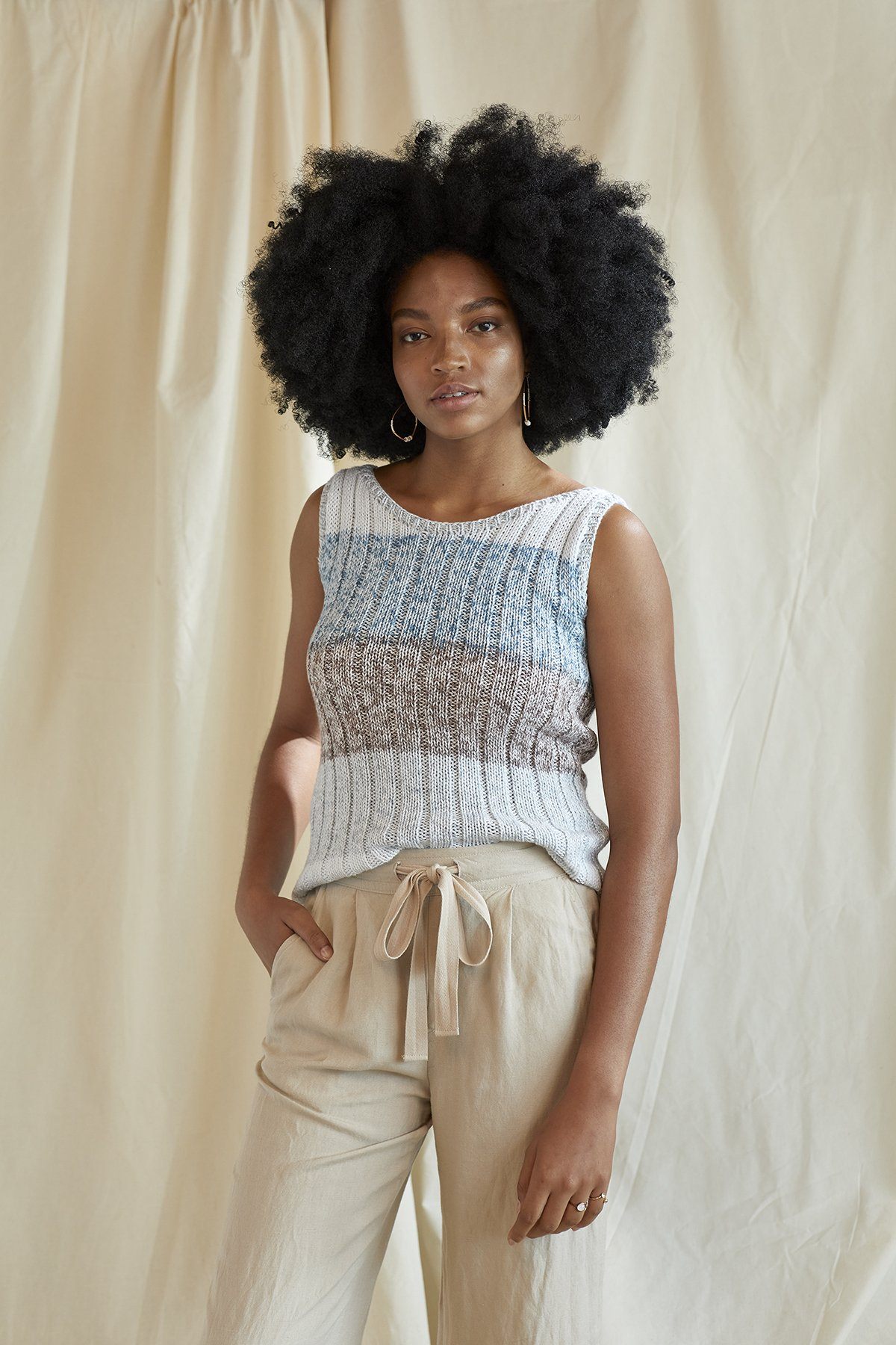 Woman wearing a sleeveless top and beige pants against a beige curtain background