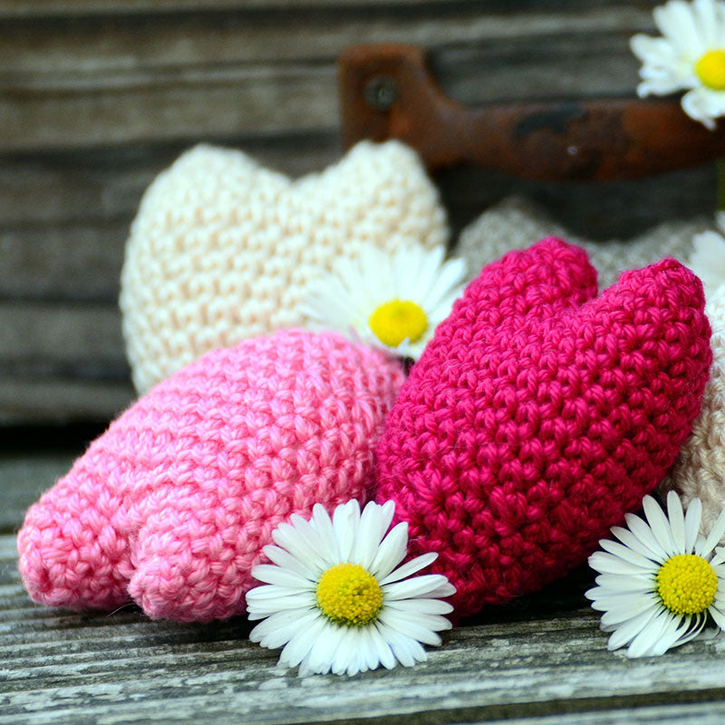 A crochet workshop featuring a pattern to create heart-shaped items, displayed with pink and red crocheted hearts and white daisy flowers on a wooden surface.