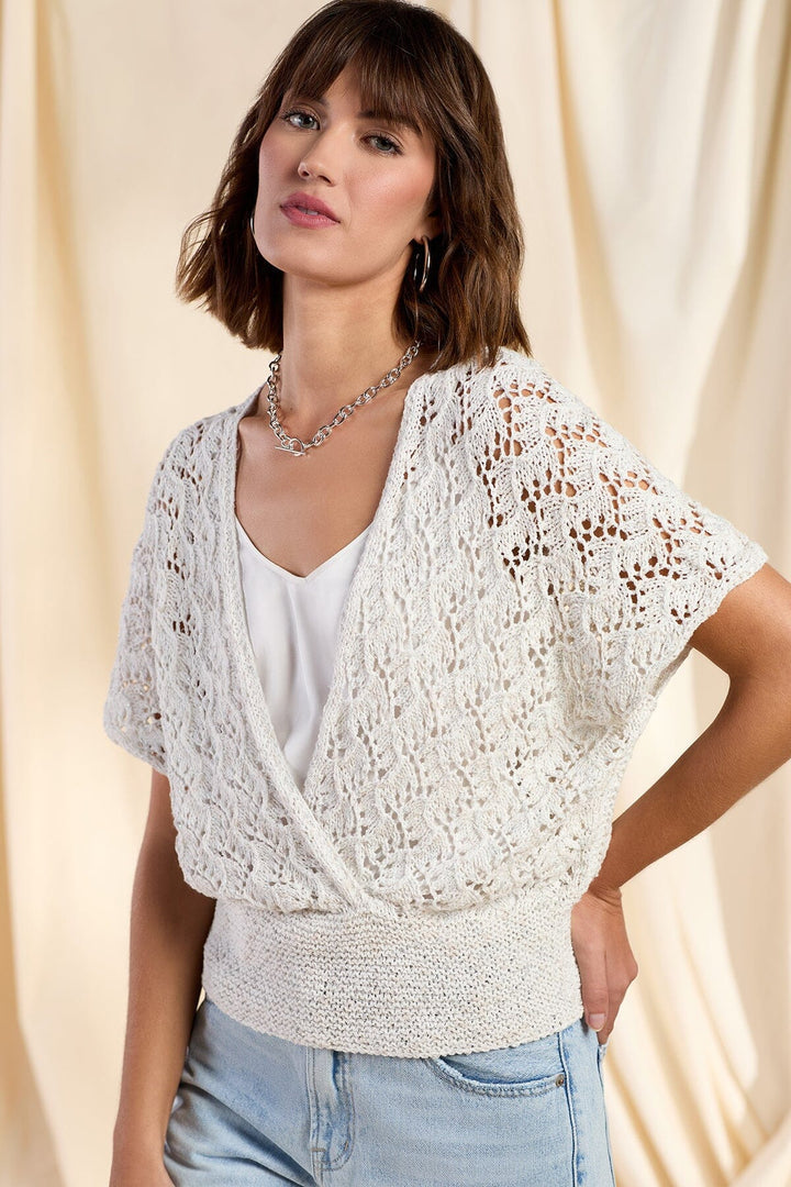 Woman wearing a white lace top over a white shirt with light blue jeans against a beige curtain background
