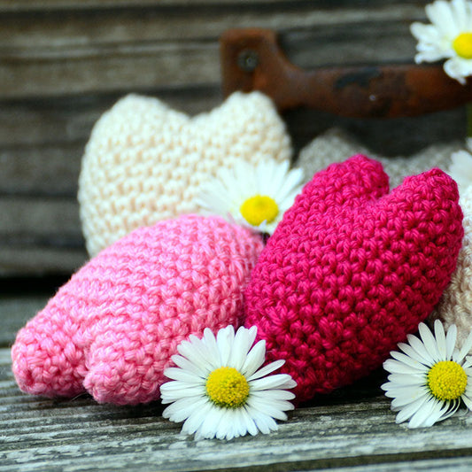 A crochet workshop featuring a pattern to create heart-shaped items, displayed with pink and red crocheted hearts and white daisy flowers on a wooden surface.